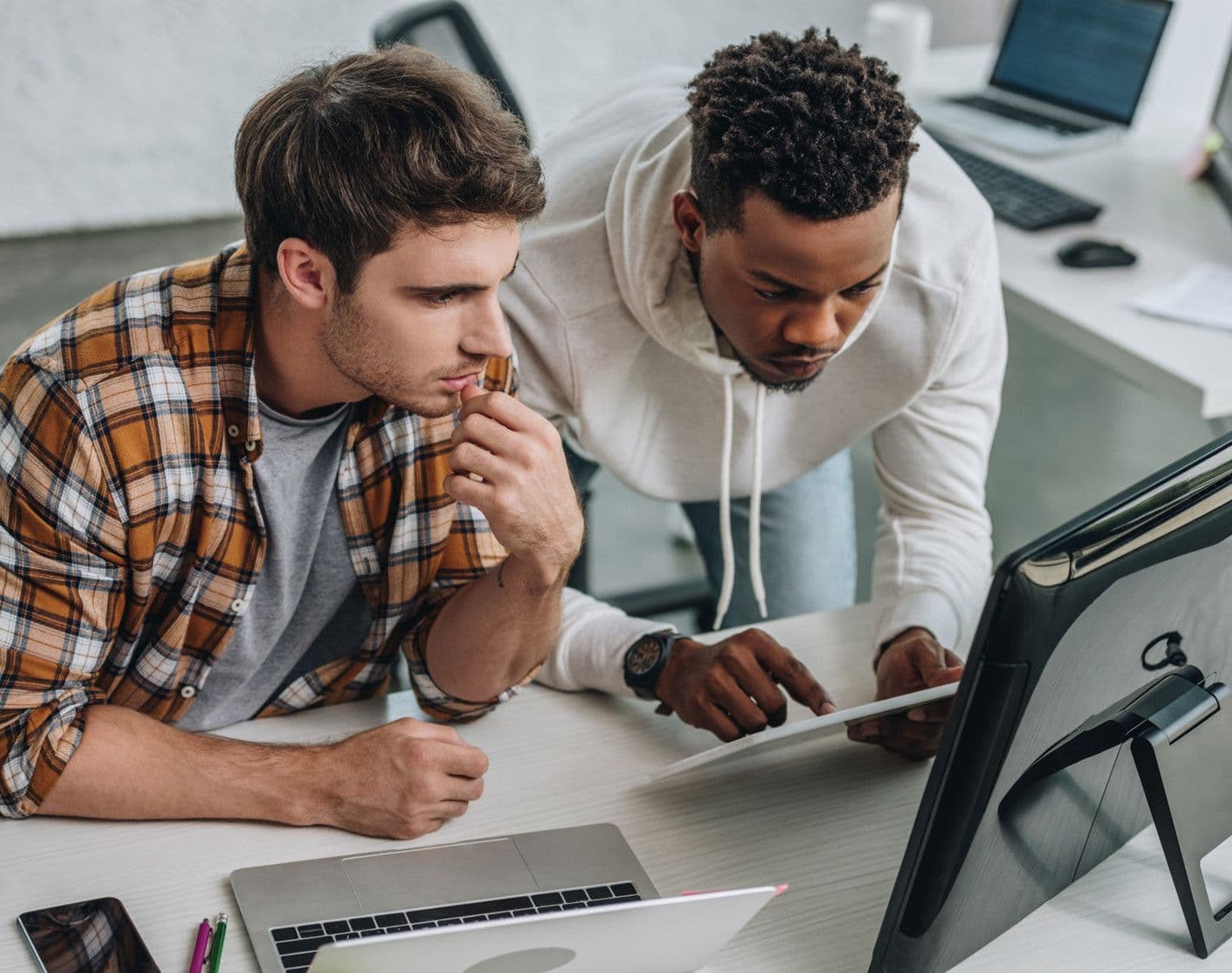 Two diverse colleagues collaborate, intently viewing a tablet and computer screen in an office.
