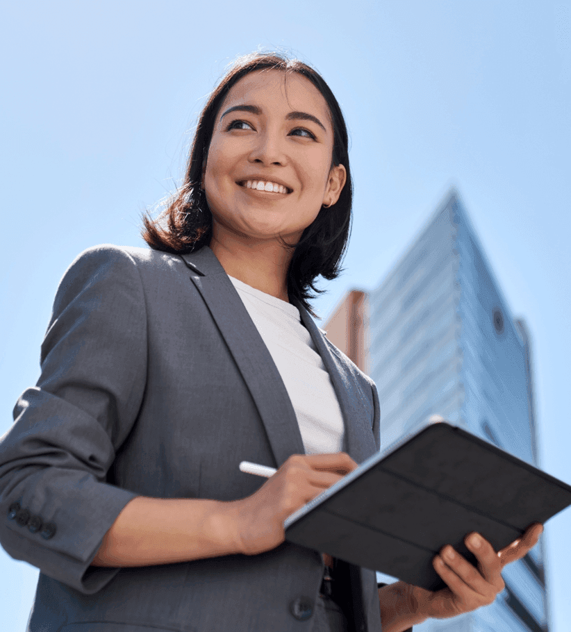 Young professional woman in a suit smiles while using a tablet outdoors.