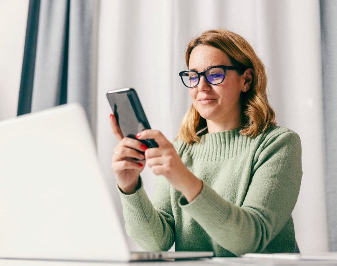 Woman in glasses smiling, holding a smartphone while looking at a laptop on a desk.