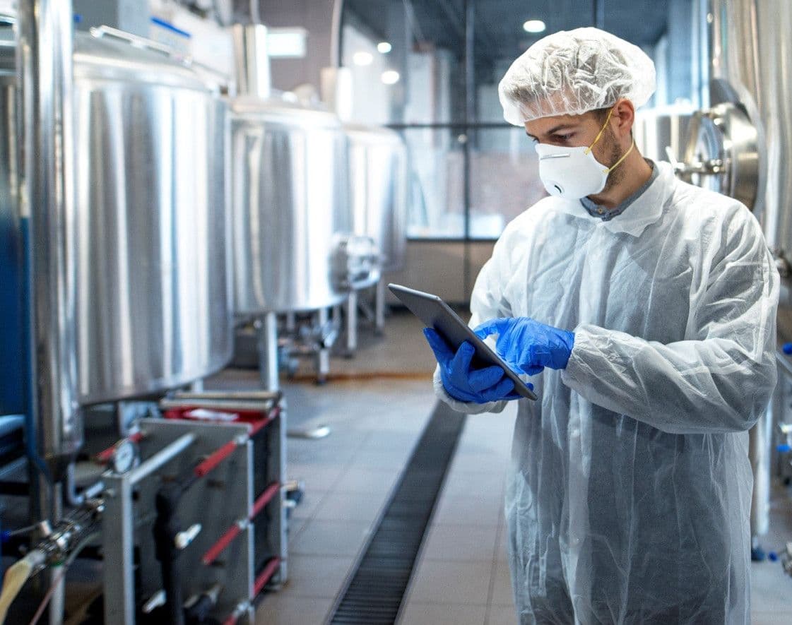 Scientist in lab coat, mask, and gloves inspecting industrial tanks with a tablet.