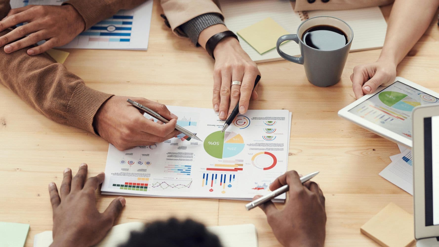 Diverse hands collaborate around a table, analyzing financial charts and data on a document.