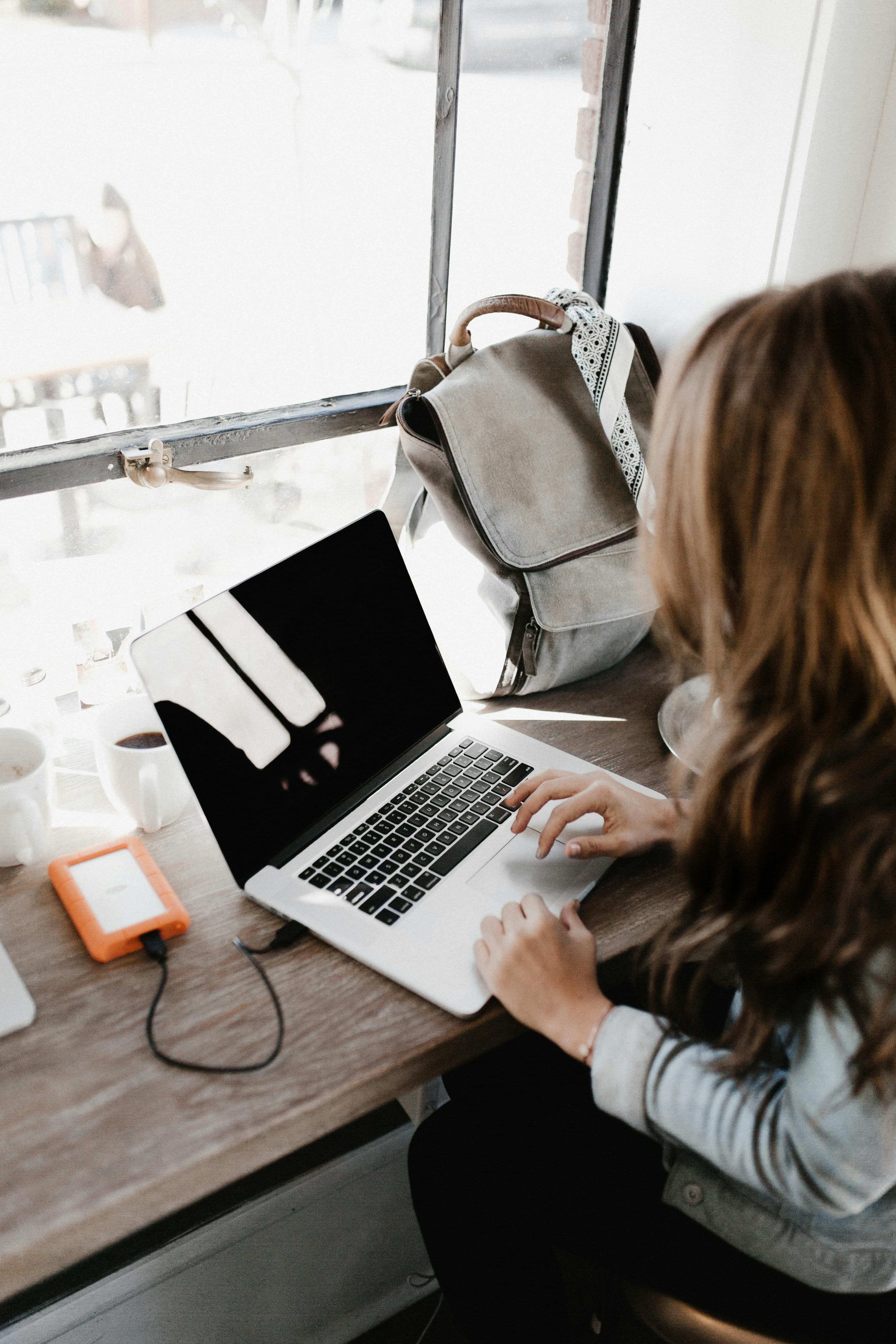 Person with long hair working on a silver laptop at a desk near a window.
